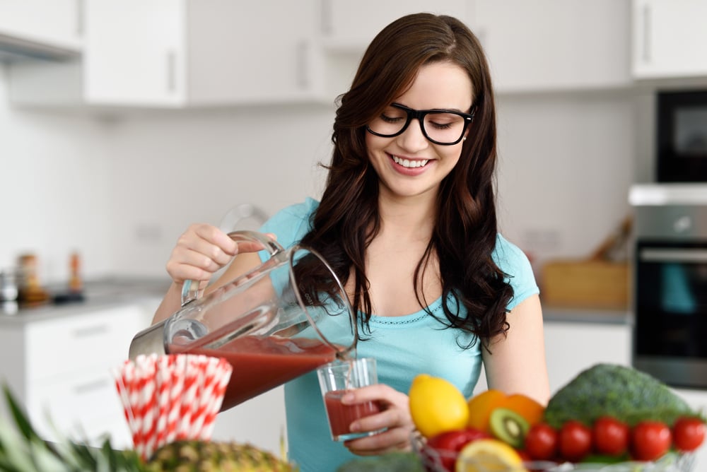 Happy healthy young woman wearing glasses pouring vegetable smoothies freshly made from assorted vegetable ingredients on her kitchen counter Happy healthy young woman wearing glasses pouring vegetable smoothies freshly made from assorted vegetable ingredients on her kitchen counter